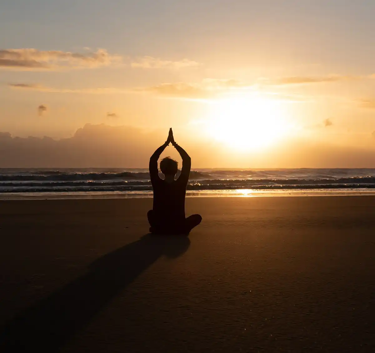A women in silhouette on a beach at sunset doing a seated yoga pose.