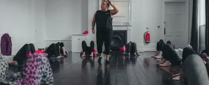 A woman stands at the front of a indoor pilates class, while women on mats excercise.