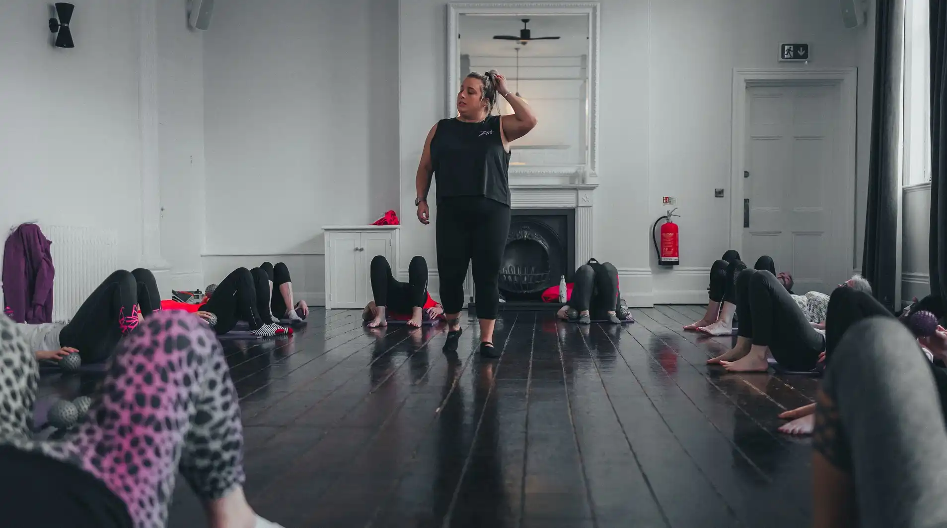 A woman stands at the front of a indoor pilates class, while women on mats excercise.