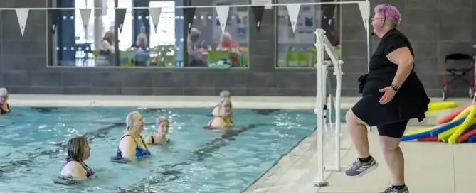 An instructor stands next to a indoor swimming pool and teaches an aquacise session.