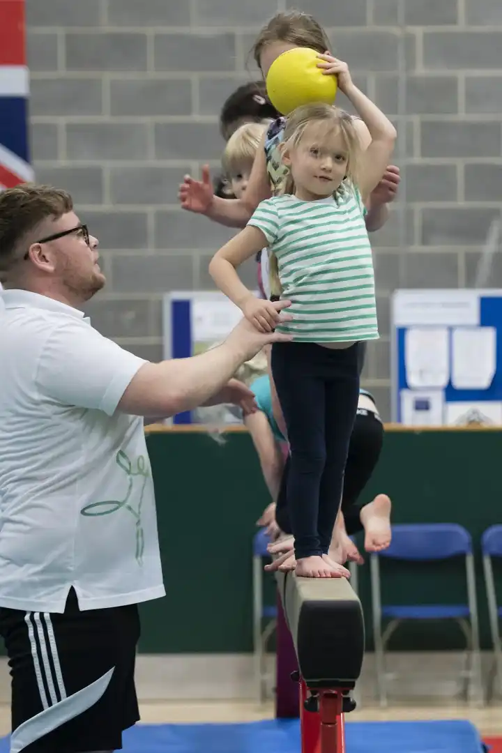 A man coaches young children walking across a beam, holding a ball.