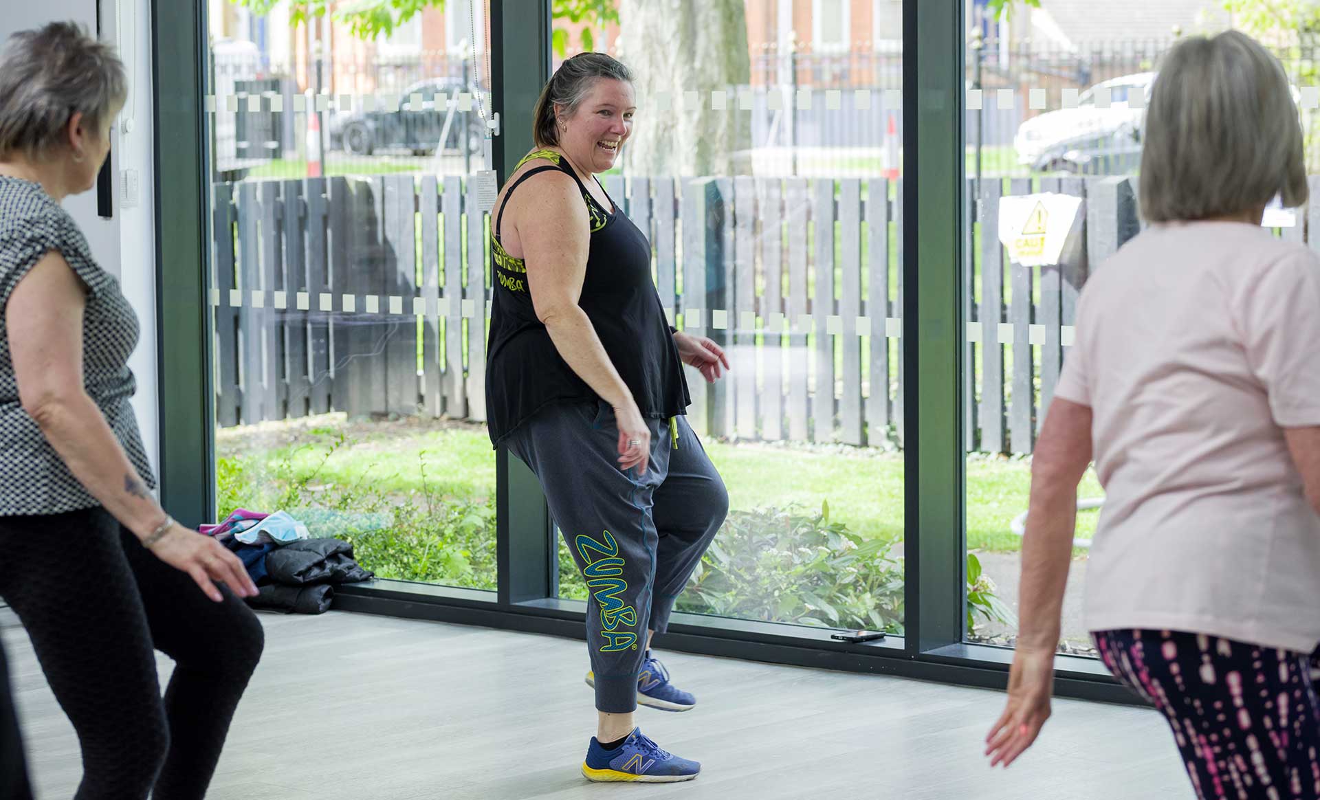 A lady in fitness clothes leads a zumba class in front of glass windows. Class participants can be seen in the foreground.