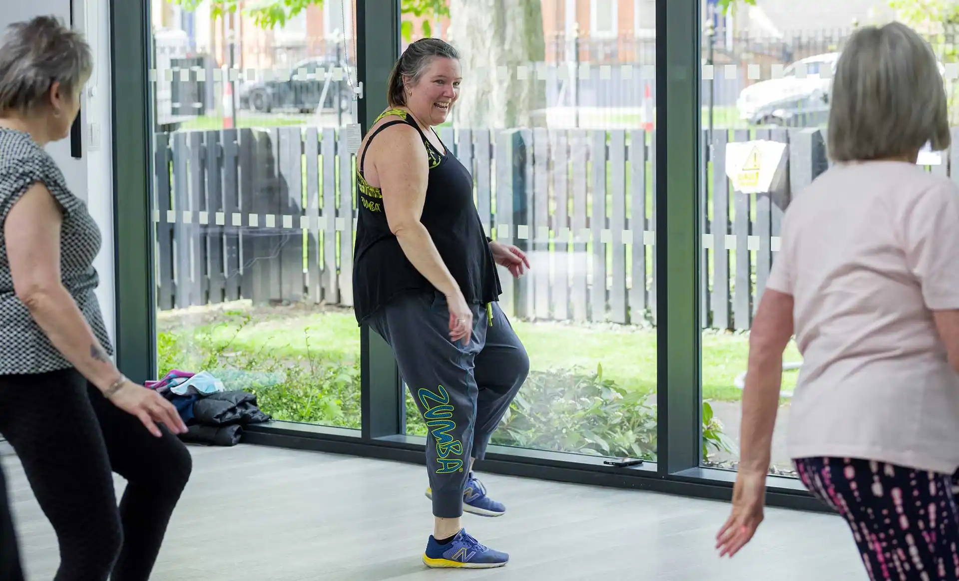 A lady in fitness clothes leads a zumba class in front of glass windows. Class participants can be seen in the foreground.