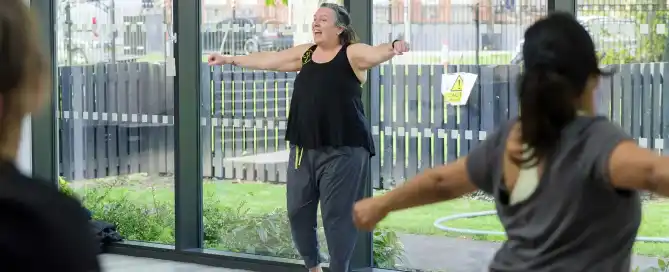 A lady stands at the front of a fitness class, with her arms outstretched, class participants seen from the back are visible in the foreground.