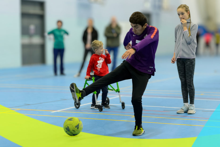 A boy in a sports hall kicks a yellow football, while a girl and boy n a walker look on.